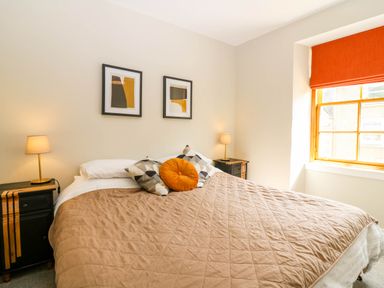 A bedroom with a bed covered by a brown quilt two bedside tables with lamps two framed pictures above the bed and a window with an orange blind at 49 Castlegate in Jedburgh