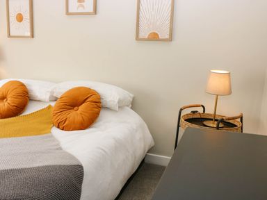 A bedroom with a bed featuring white pillows orange round cushions and a multicolored blanket next to a wicker side table with a lamp at 49 Castlegate in Jedburgh