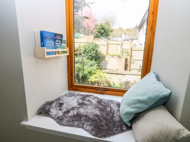 A window seat with cushions and a fur rug next to a wooden window frame and a small bookshelf with books at 49 Castlegate in Jedburgh