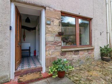 An open wooden door with patterned flooring inside a stone and stucco exterior wall with a window and potted plants outside at 49 Castlegate in Jedburgh