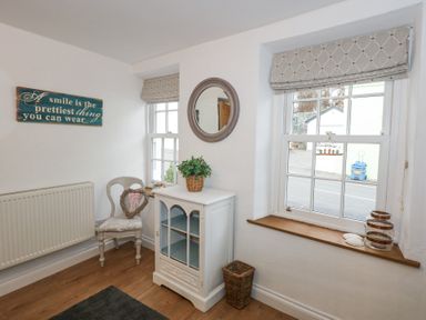 A corner of a room with a white cabinet topped with a green plant a chair with a heart-shaped wreath and two windows with Roman blinds at Pippin Cottage in Beaumaris
