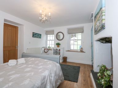 A bedroom with a bed covered with white and beige bedding two folded towels on the bed wooden door two windows with roman blinds white cabinet round mirror armchair gray rug and a fireplace with floral arrangement at Pippin Cottage in Beaumaris