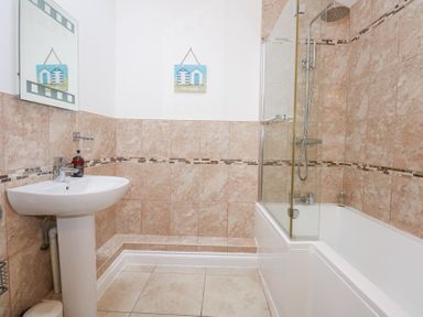 A bathroom with a pedestal sink a mirror a bathtub with a shower and tiled walls at Pippin Cottage in Beaumaris