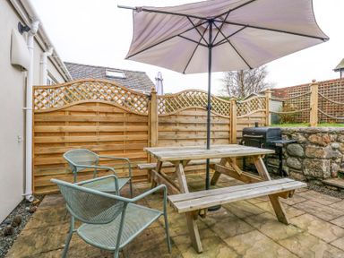 A patio with a wooden picnic table and umbrella two metal chairs wooden fence and a barbecue grill at Pippin Cottage in Beaumaris