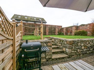 An outdoor patio area with a black barbecue grill stone steps leading to a raised lawn with wooden posts and rope fencing and a brick wall in the background at Pippin Cottage in Beaumaris