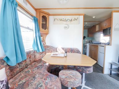 A dining area with patterned seating and a wooden table next to a window with blue curtains and a view into a kitchen at Tanyfron Holiday Caravan in Bryngwran near Gwalchmai