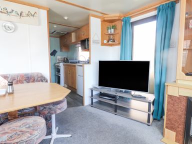 A living room with a wooden dining table and upholstered chairs a television on a stand and a view into a kitchen at Tanyfron Holiday Caravan in Bryngwran near Gwalchmai