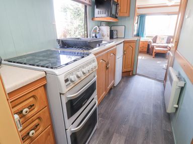 A narrow kitchen with stove sink cabinets and a view into a living room at Tanyfron Holiday Caravan in Bryngwran near Gwalchmai