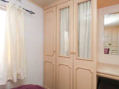 A bedroom corner with beige wardrobes with glass panels and curtains and a window with white curtains at Tanyfron Holiday Caravan in Bryngwran near Gwalchmai