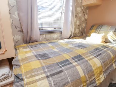 A bedroom with a bed covered by a yellow and gray plaid blanket next to a window with blinds and curtains at Tanyfron Holiday Caravan Bryngwran near Gwalchmai
