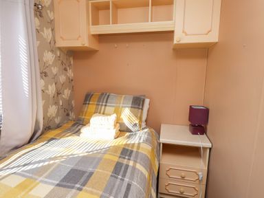 A single bed with plaid bedding and folded towels next to a nightstand with a lamp in a small bedroom at Tanyfron Holiday Caravan in Bryngwran near Gwalchmai