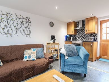 A living room with a brown sofa and blue armchair next to a kitchen with wooden cabinets at Oak Cottage in Aylesbeare