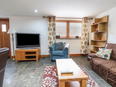 A living room with a television on a wooden stand a blue armchair a brown sofa a wooden coffee table and shelving at Oak Cottage in Aylesbeare