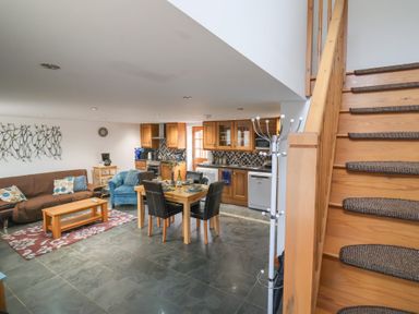 An open plan living room and kitchen with a dining table four chairs a brown sofa an armchair and wooden stairs at Oak Cottage in Aylesbeare