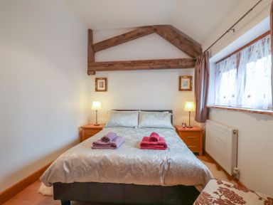 A bedroom with a double bed two bedside tables and lamps wooden beams on the wall and window with curtains at Oak Cottage in Aylesbeare