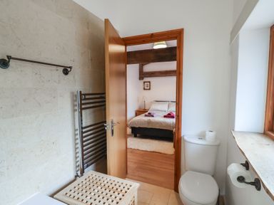 A bathroom with a toilet, towel rack, and a doorway leading to a bedroom with a bed and wooden beams at Oak Cottage in Aylesbeare