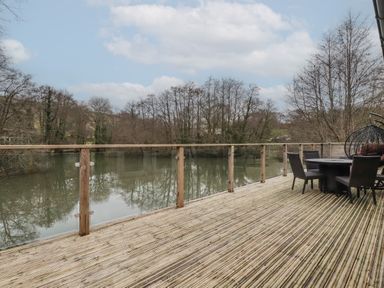 A wooden deck with glass railing overlooking a river with trees and outdoor table and chairs at 2 Millers Island Near Lanreath