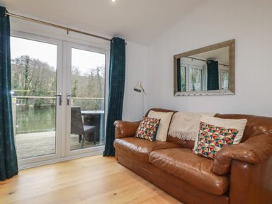 A living room with a brown leather sofa, patterned cushions, a floor lamp, glass doors leading to a balcony overlooking a body of water at 2 Miller's Island Near Lanreath