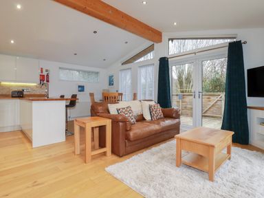 A living room with a brown leather sofa a wooden coffee table and side table a white rug and kitchen area at 2 Miller's Island Near Lanreath