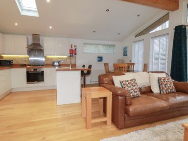 An open kitchen and living room with a brown leather sofa wooden dining table bar stools and white cabinets at 2 Miller's Island Near Lanreath