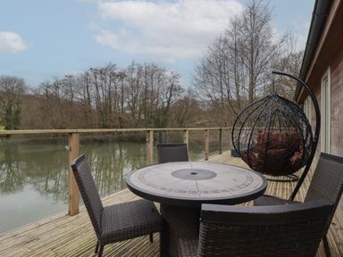An outdoor deck with a round table surrounded by four chairs and a hanging egg chair overlooking a river at 2 Miller's Island Near Lanreath
