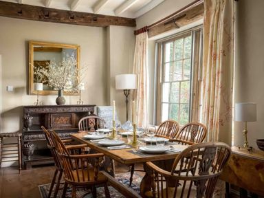 A dining room with wooden table and chairs set near a large window with curtains and an ornate sideboard with a mirror