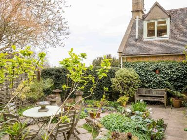 A garden with wooden chairs and a table surrounded by plants and a stone house in the background at Courtyard House Blockley