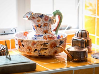 A floral patterned ceramic pitcher inside a matching basin on a yellow tiled countertop with a metal watering can and other items at Courtyard House in Blockley