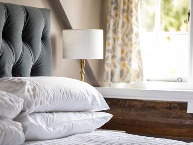 A bedroom with a bed with white pillows and a gray headboard a lamp a window with patterned curtains and wooden paneling at Courtyard House Blockley