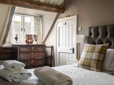 A bedroom with a bed with folded towels a wooden dresser with a copper pitcher and a window with curtains at Courtyard House in Blockley