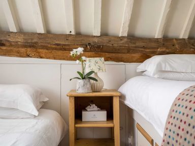 A wooden nightstand with a lamp and potted orchid between two beds with white bedding and exposed wooden beam on the wall at Courtyard House in Blockley