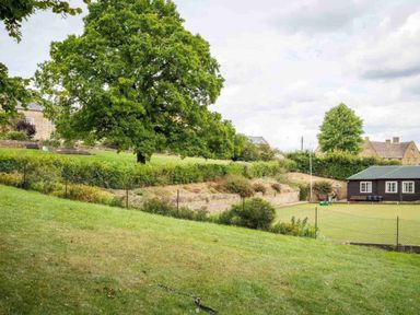 A garden with green grass a large tree bushes and a dark wooden shed near houses at Courtyard House in Blockley