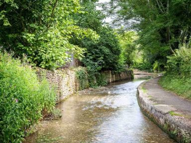 A narrow river bordered by a stone wall and path surrounded by green trees and plants at Courtyard House in Blockley