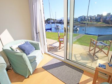 A room with a green armchair and blue cushions next to glass sliding doors opening to a patio with wooden chairs and a water view at Harbour Escape in Porthmadog