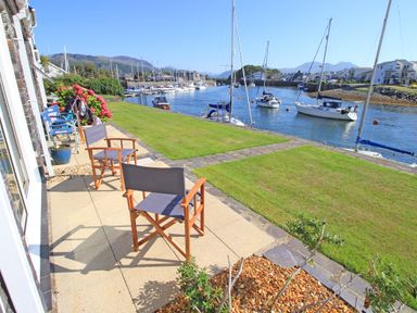 A patio with chairs and a lawn overlooking a river with sailboats and buildings in the background at Harbour Escape in Porthmadog