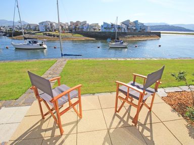 Two wooden chairs with fabric seats on a paved patio facing a river with sailboats and modern houses on the opposite shore at Harbour Escape in Porthmadog