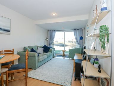 A living room with green sofa blue cushions wooden dining table and chairs shelving unit and glass doors with curtains at Harbour Escape in Porthmadog