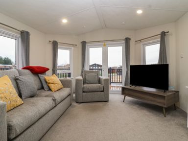 A living room with a grey sofa and armchair a wooden TV stand and curtains at Longdon Lodge in Wickhamford near Evesham