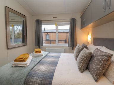A bedroom with a bed with patterned pillows folded towels on the bed a window with curtains and a wall mirror at Longdon Lodge in Wickhamford near Evesham