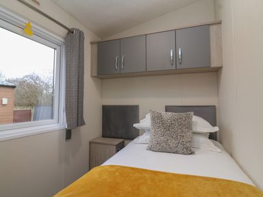 A small bedroom with a single bed a yellow blanket a gray pillow overhead cabinets and a window with a curtain at Longdon Lodge in Wickhamford near Evesham