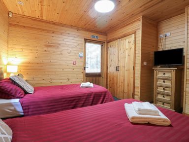 A bedroom with two single beds with red striped bedding a wooden wardrobe a TV on a wooden chest of drawers and wooden walls and ceiling at Rowan Lodge in Rosliston