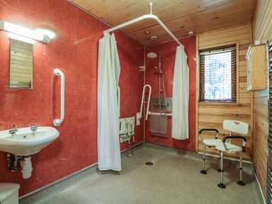 A bathroom with a wall-mounted sink a shower area with white curtains a shower chair and wooden wall panels at Rowan Lodge in Rosliston
