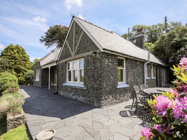 An exterior view of a stone house with a slate roof and a patio with outdoor table and chairs surrounded by plants and trees at Bryn Awelon in Harlech