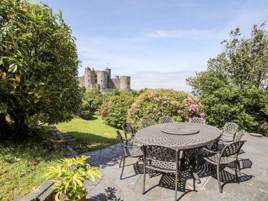 An outdoor patio with a round metal table and eight chairs surrounded by green bushes with a castle in the background at Bryn Awelon in Harlech