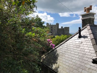 A stone castle in the distance surrounded by green trees next to a house roof with chimney stacks in Harlech