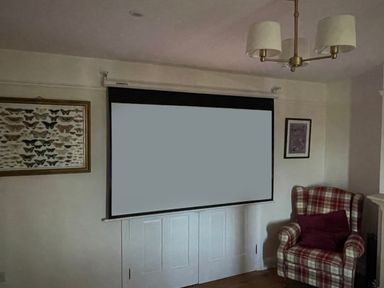 A living room with a projector screen hanging over white double doors a framed butterfly picture on the left wall a framed picture on the right wall a plaid armchair with a cushion and a ceiling light fixture at Bryn Awelon in Harlech