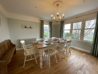 A dining room with a wooden table set with plates and bowls surrounded by white chairs and a wooden bench near windows with curtains at Bryn Awelon in Harlech