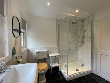 A bathroom with a sink, toilet with wooden seat, glass-enclosed shower, small white table, and a round mirror at Bryn Awelon in Harlech
