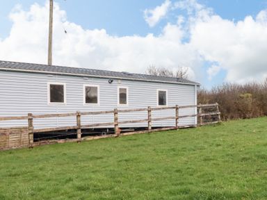 An exterior view of a light blue caravan with four windows and a wooden railing on a grassy hill at Little Trevorrick Caravan in St Issey