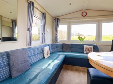 A seating area with blue cushioned benches and a round wooden table by windows with striped curtains and flowers at Little Trevorrick Caravan in St Issey